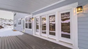 Covered deck with white ceiling, wall lights, and large windows along the back of the home